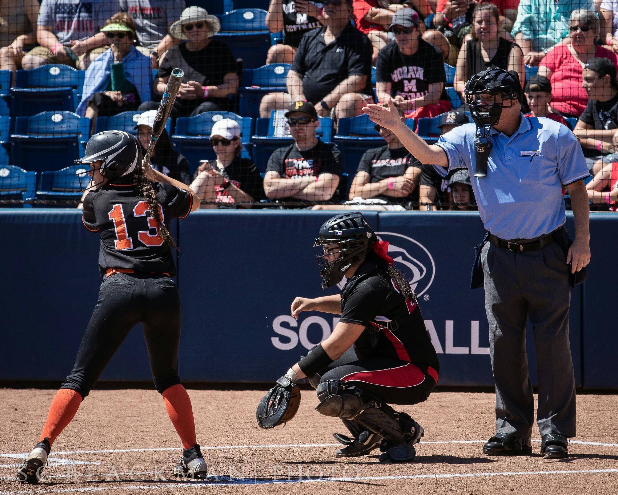 softball player on left preparing to swing bat with catcher to her right kneeling and the umpire further right pointing with his hand