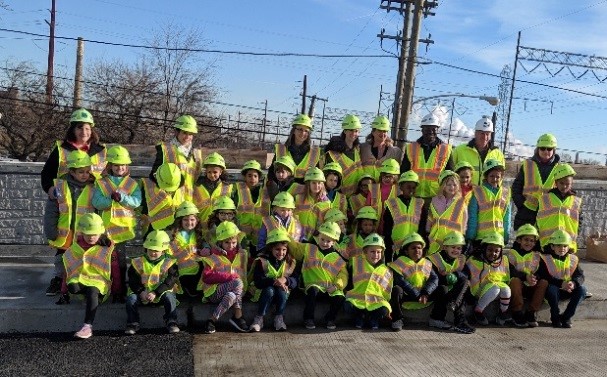 Large group of elementary students wearing bright yellow construction vests and hard hats.