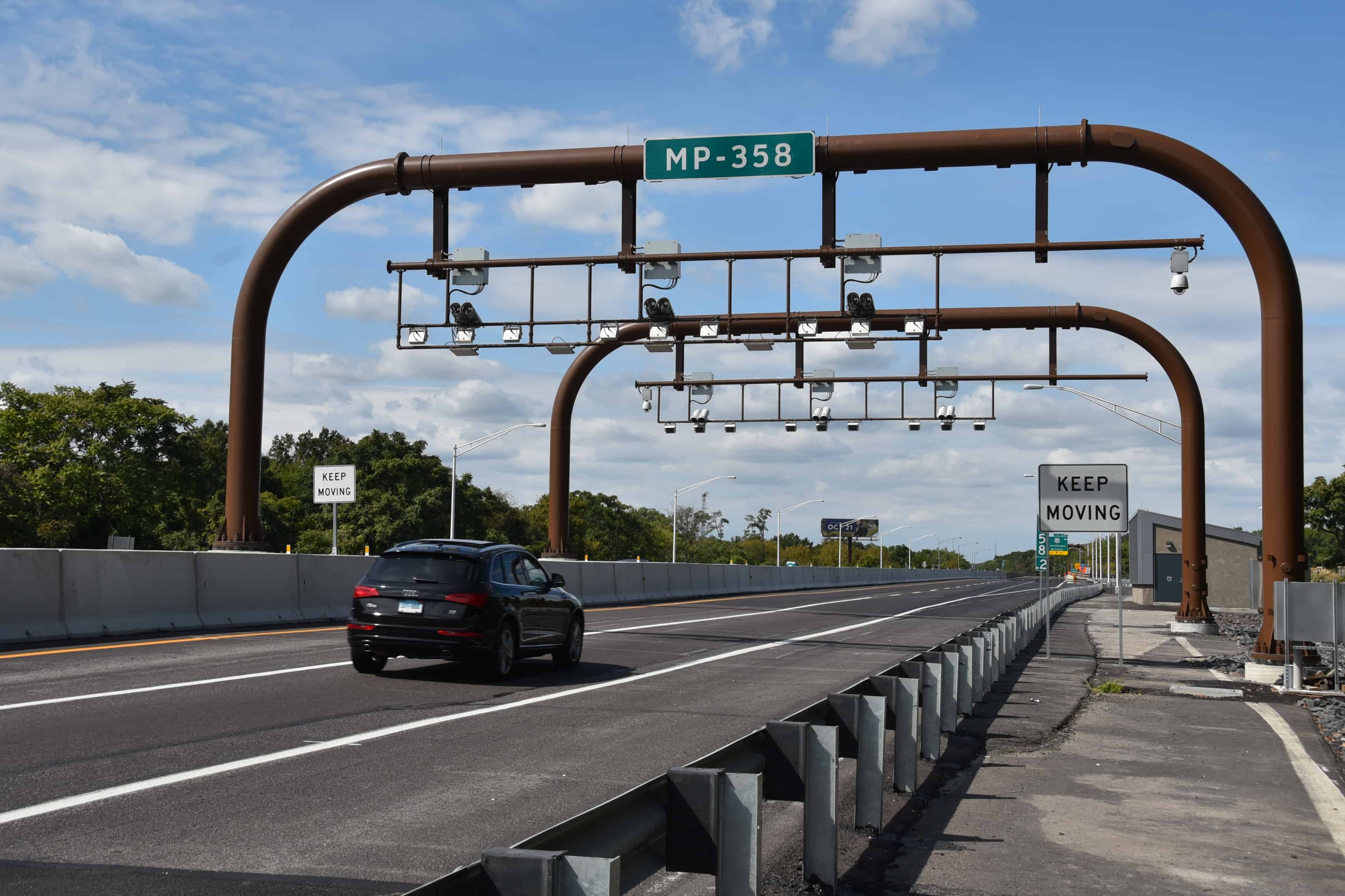 A black SUV drives under two steel gantries with electronic tolling cameras mounted to them. A sign to the right of the road says 'Keep Moving'.