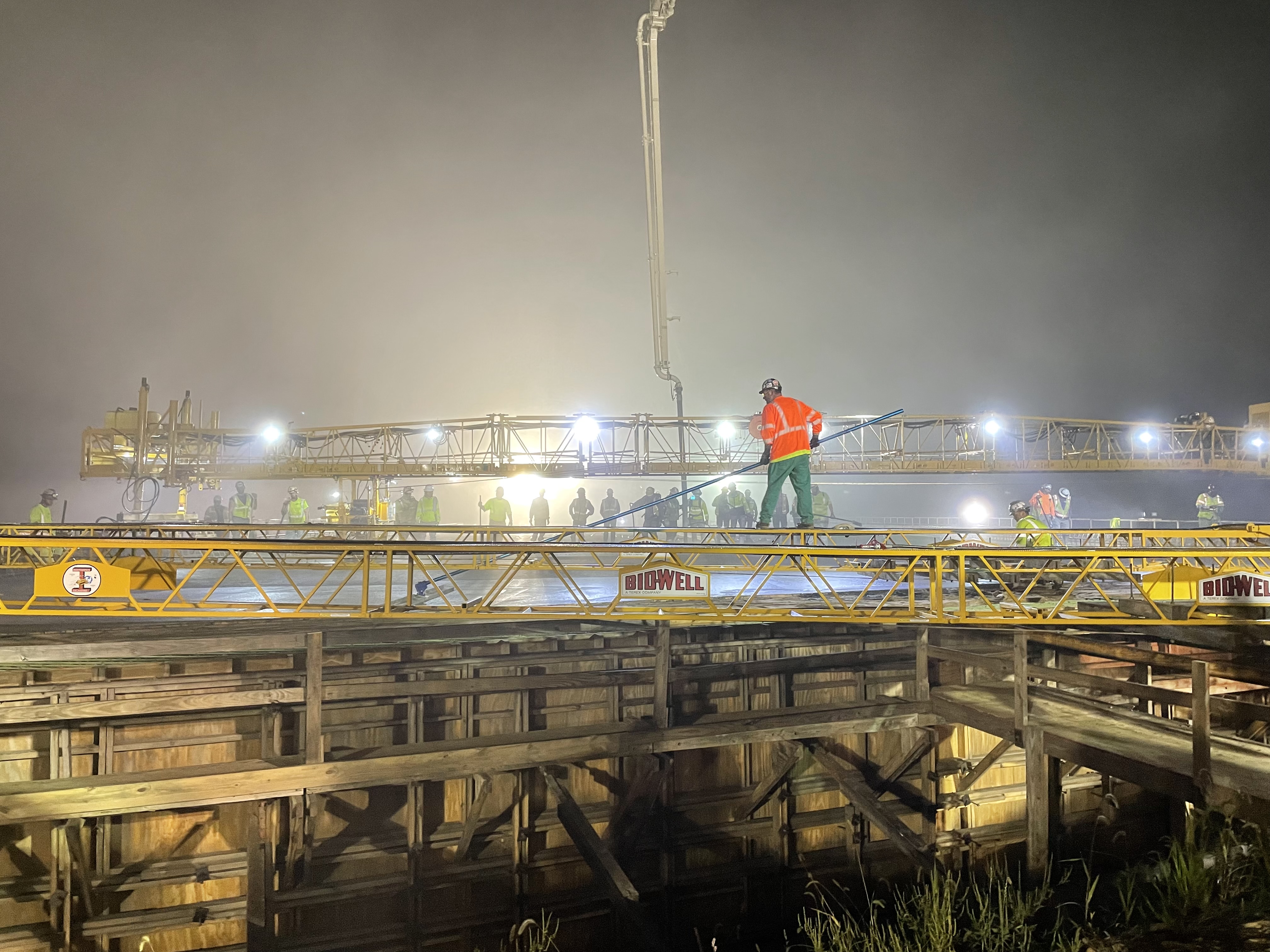 Construction workers in safety gear work on top of a bridge at night.