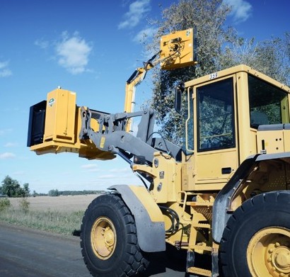 Heavy equipment using a brushing loader attachment to trim a tree along a road.