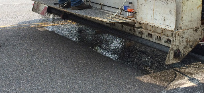 Truck on a road sprinkling dark stone-like aggregate onto a wet surface.