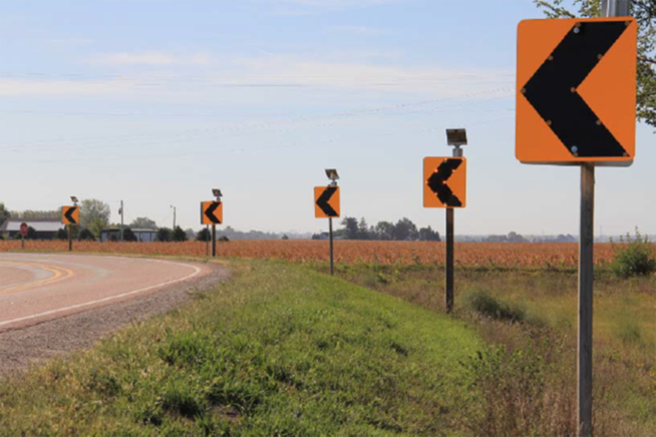 Orange and black signs pointing left along a curve of the roadway in a rural setting.