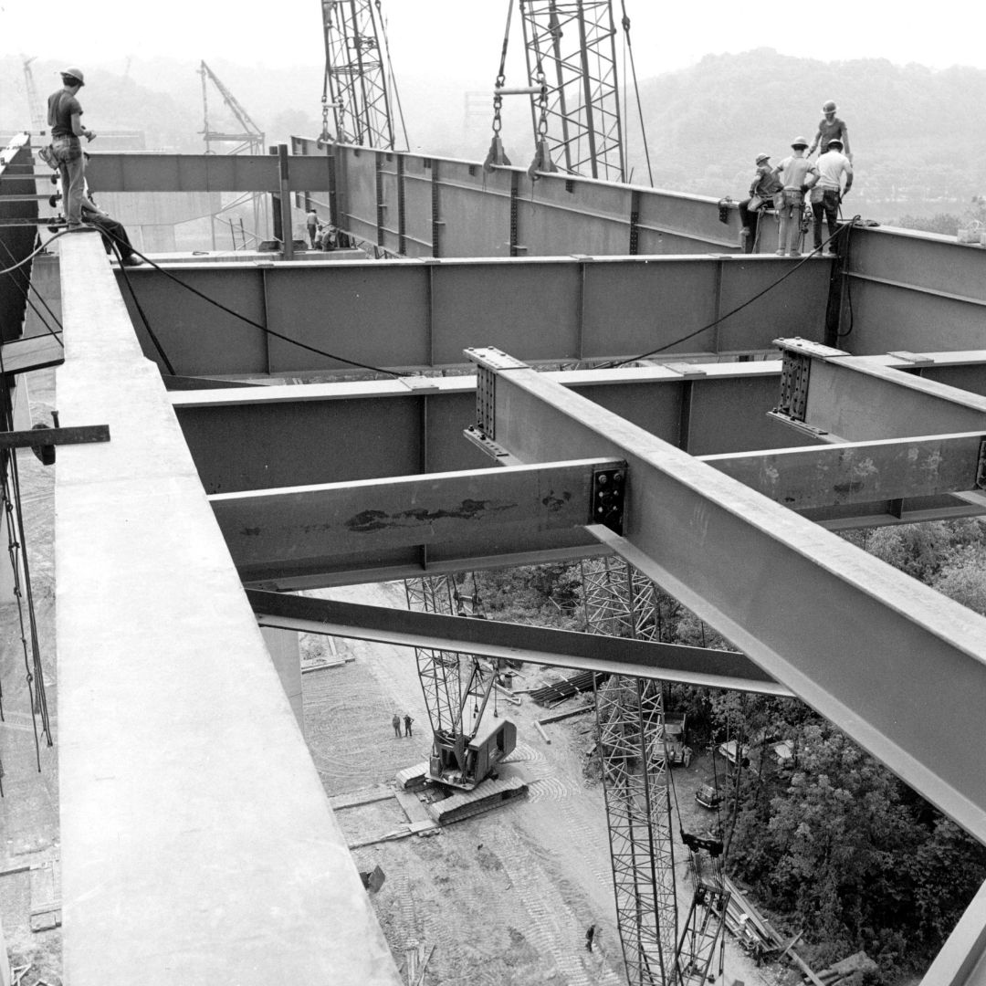 Black-and-white photo of workers standing on top of metal bridge beams high above the ground.