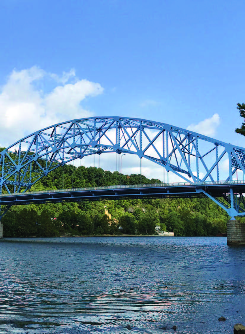 Side view of a blue steel bridge stretching across the Monongahela River with tree-covered hills in the background.