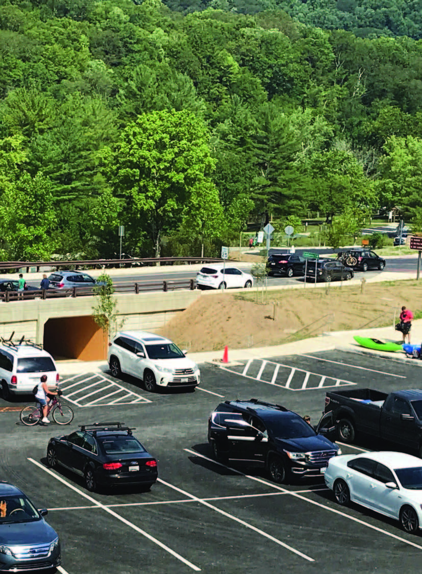 Vehicles are parked in a lower lot, one person on a bike, another preparing a kayak, as more vehicles cross a bridge above the parking lot with trees in the background.