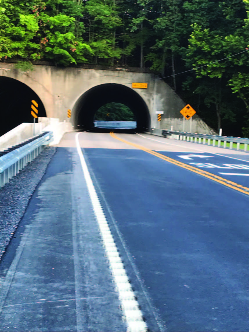 A freshly paved two-lane road goes through a concrete bridge tunnel topped with trees.