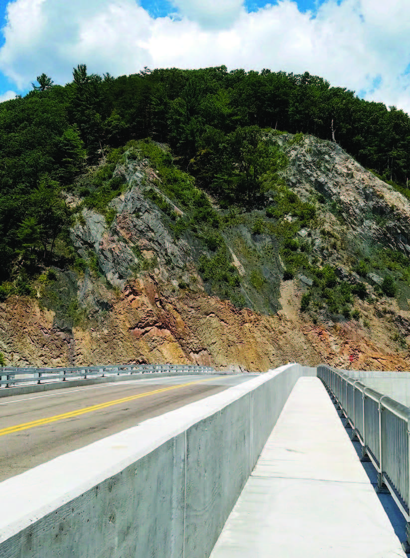 A two-lane road crosses a concrete bridge with a pedestrian walkway to the right and a tree-topped mountain in the background.