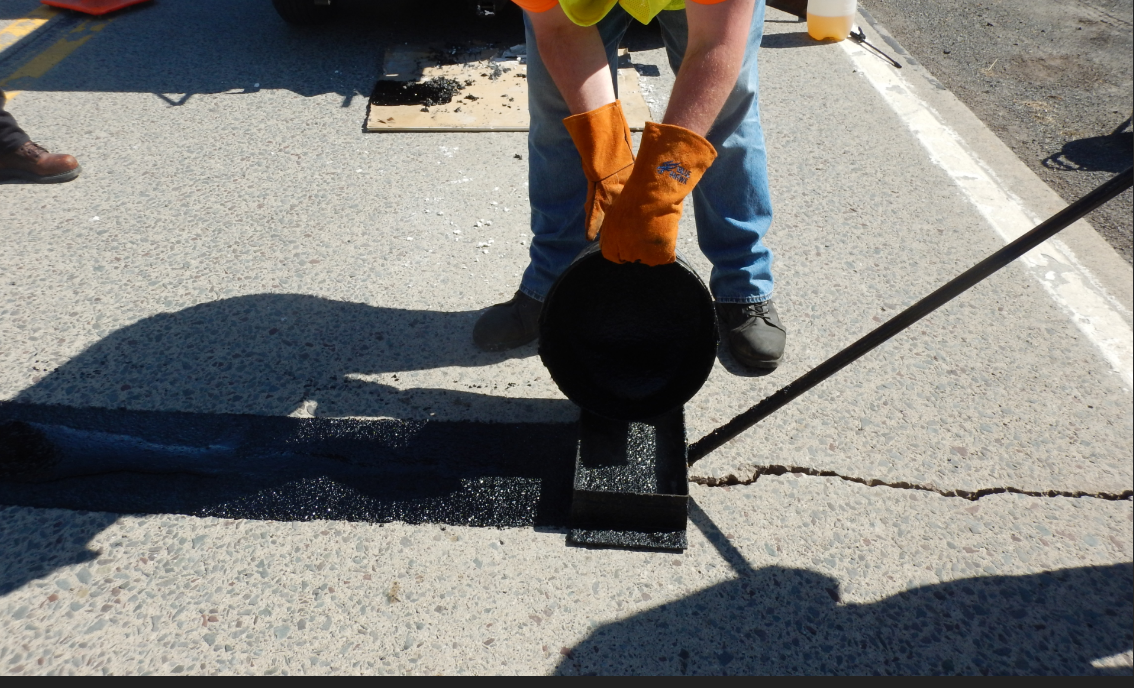 A worker with heavy orange gloves pours hot pour mastic material from a bucket through a rectangular frame onto a crack in the pavement.