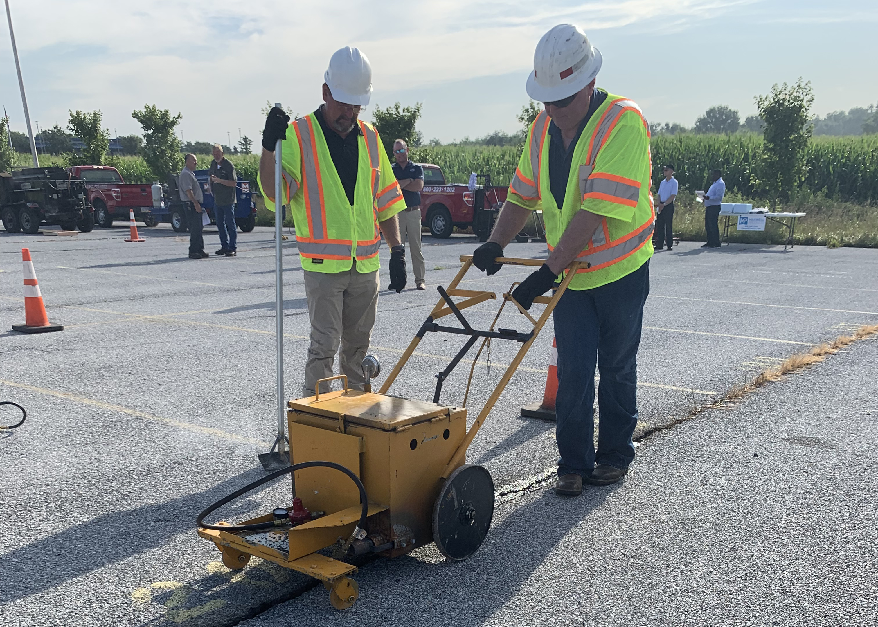 Two workers in yellow construction vests and white construction helmets use a hot pour mastics machine to fill a crack in pavement.