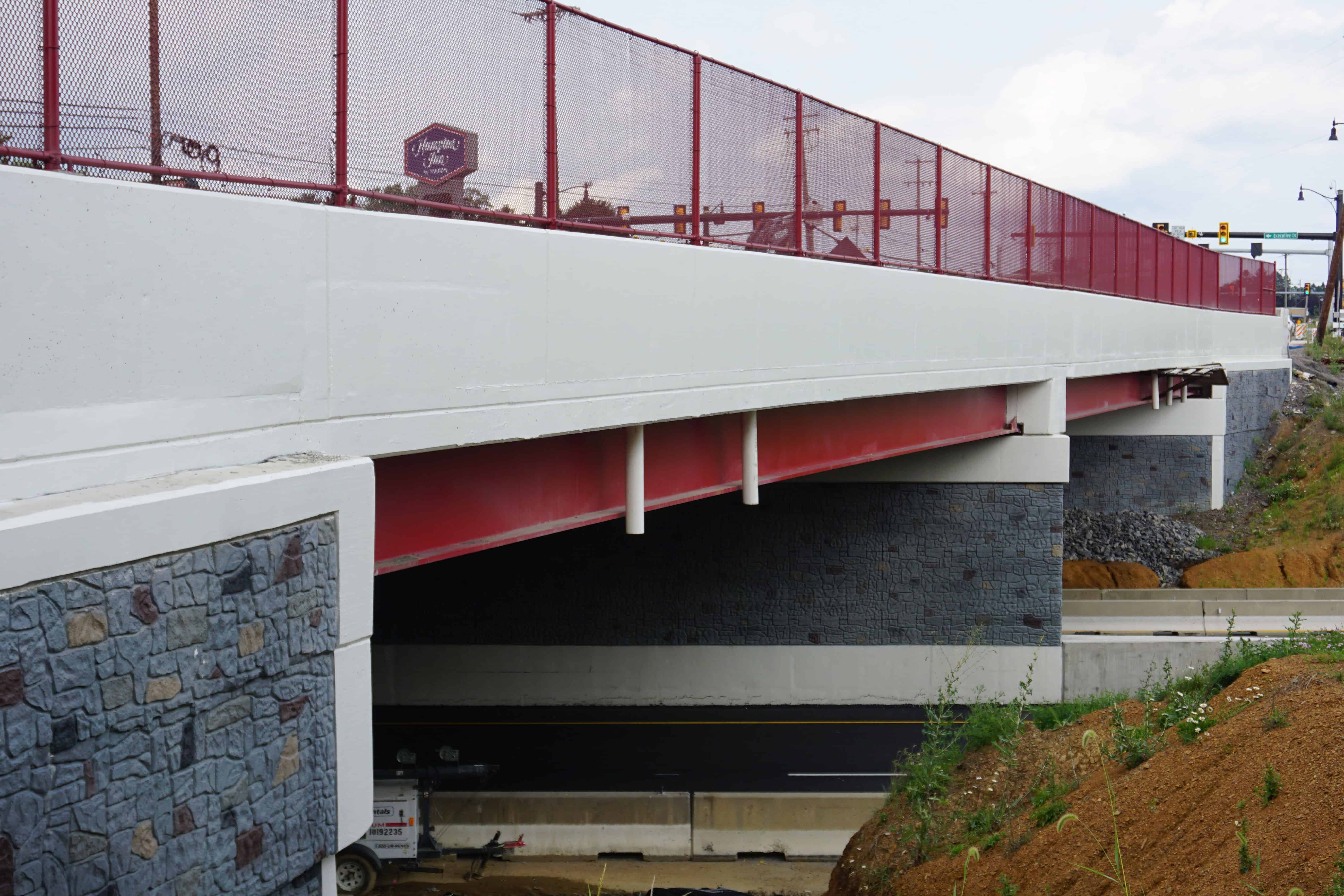 A concrete bridge with red steel beams and fencing stretches across two roadways.