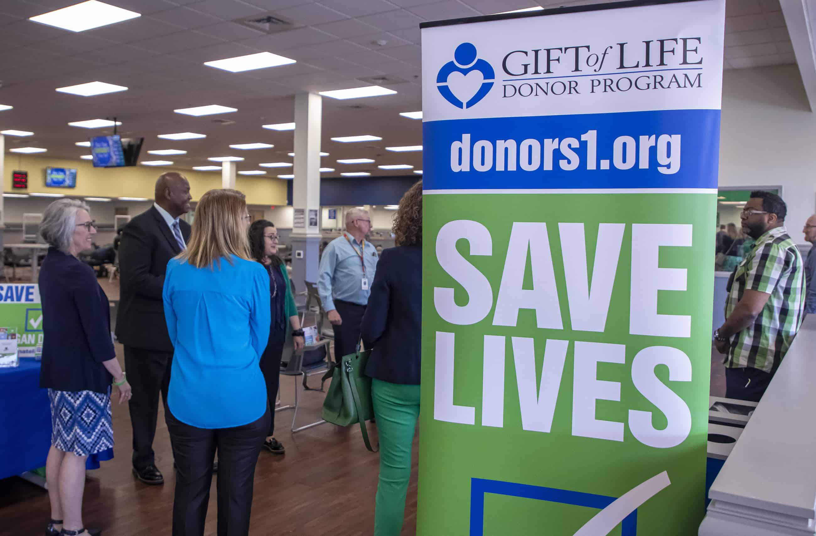 A group of people stand to the left of a large banner that reads Save Lives and promotes the Gift of Life Donor Program.